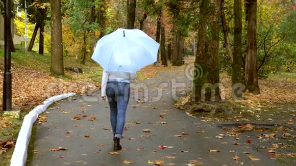 穿着白色皮夹克的女人带着蓝色雨伞在秋天公园散步视频的预览图