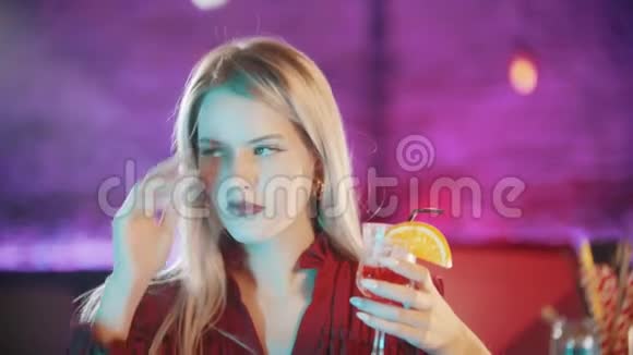 漂亮的金发女郎涂着红色口红坐在酒保的旁边玩着她的头发霓虹灯蓝色视频的预览图