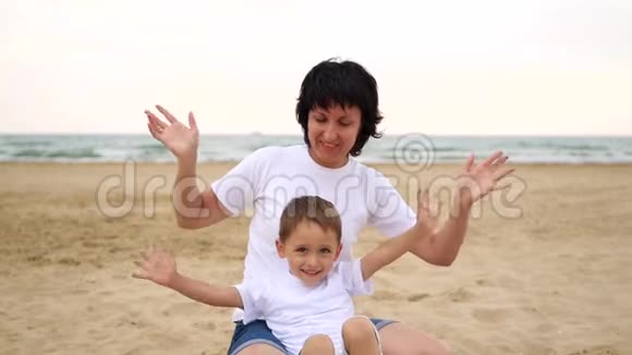 一位快乐的母亲和她的孩子正微笑着双手朝坐在沙滩上的相机挥手视频的预览图
