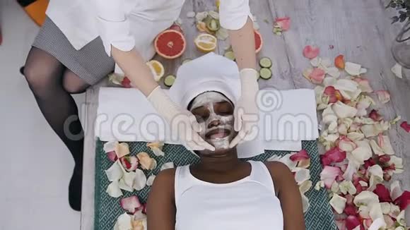 年轻的非洲裔美国妇女头上戴白毛巾躺在沙发上放松而美容医生正在应用视频的预览图