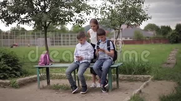 两个男孩和女孩在放学后使用他们的电话可爱的男孩坐在长凳上玩网络游戏女孩的样子视频的预览图