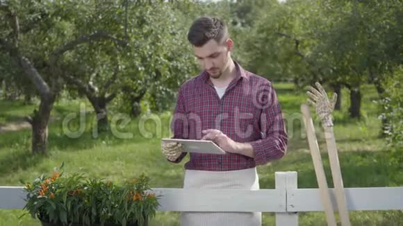 年轻的农民戴着花园手套用平板电脑站在前面的篱笆后面夏天的花园在视频的预览图
