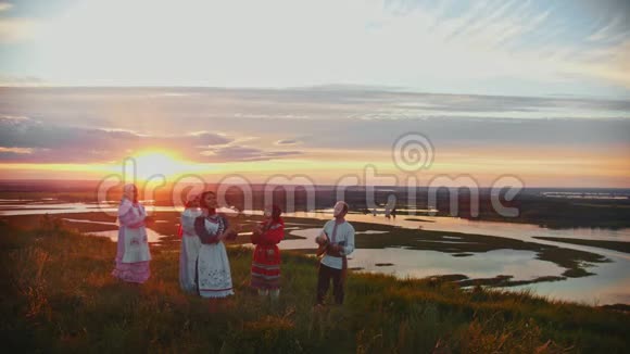 穿着俄罗斯传统服装的年轻人站在美丽的夕阳下的田野上唱歌视频的预览图