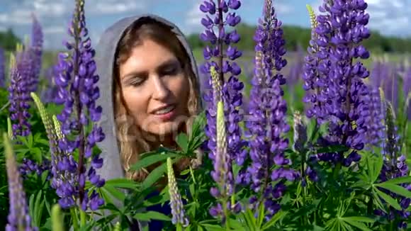 一个美丽的女人在阳光明媚的日子里欣赏着草地上的紫色花朵微笑着脸和鲜花特写视频的预览图