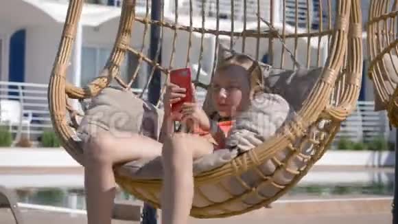 年轻女孩在度假酒店的挂椅上看着手机少女在茧椅上荡秋千视频的预览图