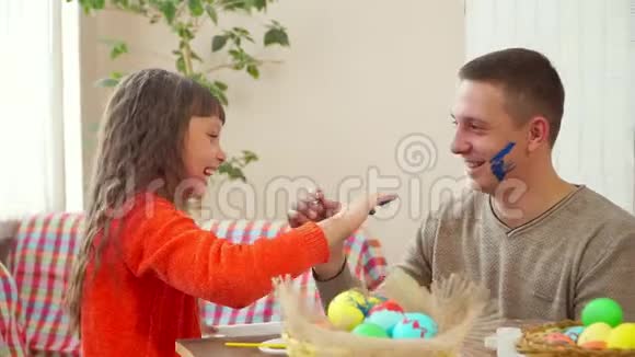 爸爸和女儿用蓝色颜料在对方的脸上涂彩蛋桌子上有一个篮子里面有复活节彩蛋视频的预览图