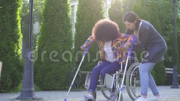 有非洲发型的非洲妇女在康复过程中在轮椅上残疾在帮助下学会走路视频的预览图