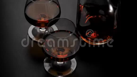 豪华白兰地带有金色干邑的圆形瓶子和两个玻璃杯在黑色背景下的黑色桌子上旋转玻璃闪闪发光视频的预览图