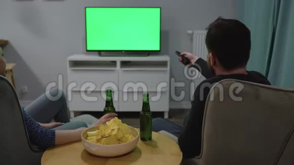 男人和女人坐在椅子上用绿色的屏幕看电视喝啤酒吃薯片后景铬键视频的预览图