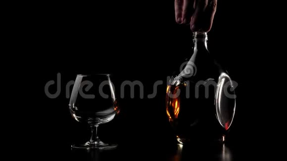 豪华白兰地手在黑色桌子上的一个玻璃旁边在黑色背景下转动一瓶金色干邑玻璃闪闪发光视频的预览图