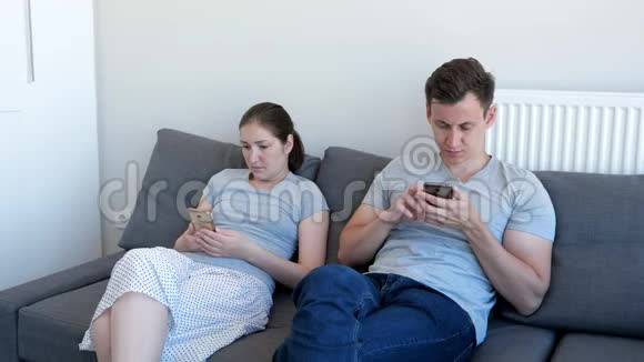 年轻夫妇男人和女人坐在沙发上看着他们的手机对小工具的依赖视频的预览图