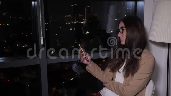 女士浏览电话坐在窗口附近晚上可以看到全景城市景色视频的预览图