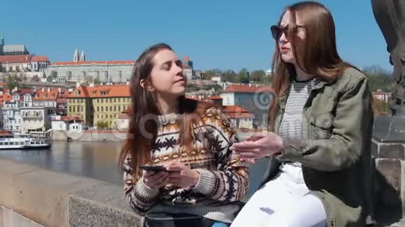 两个女人坐在桥上在布拉格市中心的背景下交谈一艘船经过女人回头看视频的预览图