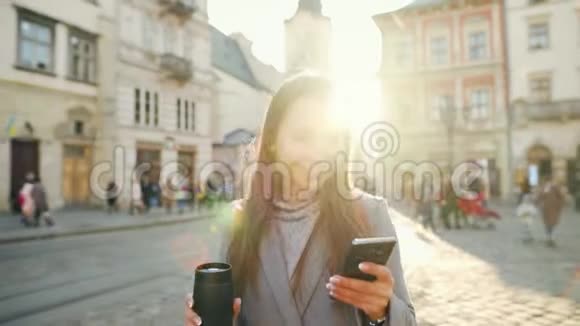 年轻的女人手里拿着一个保温杯在日落时用智能手机沿着一条老街走着交流社会视频的预览图
