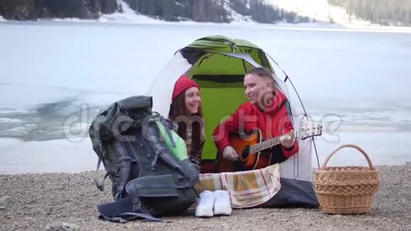 年轻可爱的夫妇坐在帐篷里在群山的背景下用吉他唱歌视频的预览图