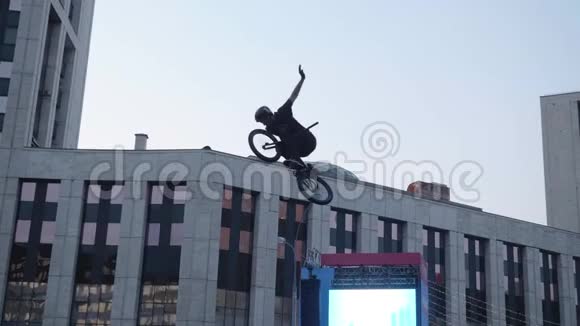 穿着黑色衣服的Biker在他的BMX自行车上跳得很高而且不用手就变魔术有窗户的现代城市建筑视频的预览图