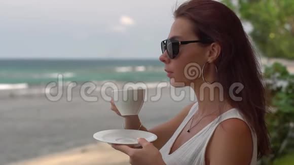 可爱的女孩戴着黑色眼镜穿着白色连衣裙在海洋上喝着一杯卡布奇诺欣赏着美好的早晨景色视频的预览图