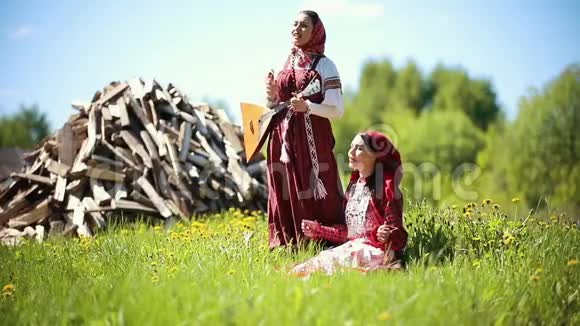 两个穿着俄罗斯传统服装的年轻女子在木柴背景下唱着巴拉莱卡的歌视频的预览图