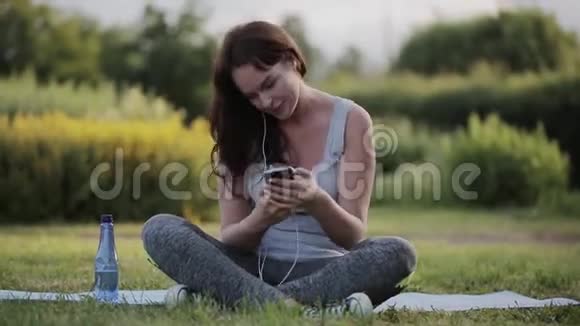 美丽的女人在训练后喝水用智能手机听音乐视频的预览图