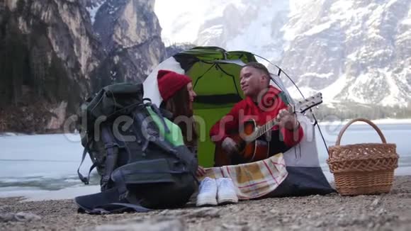 年轻夫妇在旅行坐在帐篷里在群山的背景下用吉他唱歌视频的预览图