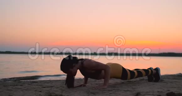 运动女孩在日出时在海滩上做俯卧撑穿着运动上衣和腿做俯卧撑的黝黑的年轻微笑女孩视频的预览图