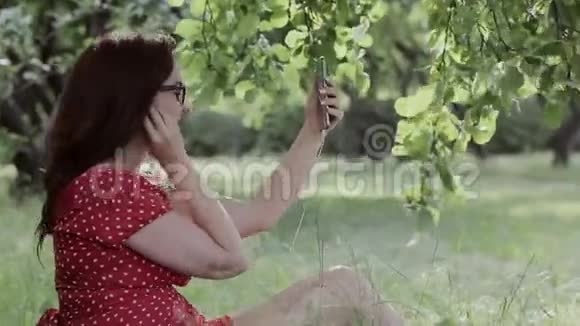超大号女孩坐在树下的草地上用智能手机听音乐视频的预览图