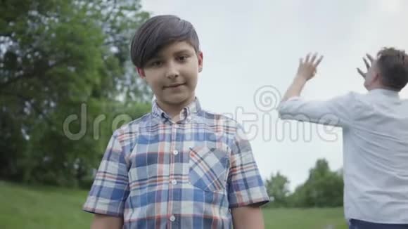 可爱的男孩看着镜头站在前台而他的爸爸或哥哥在踢足球投掷视频的预览图
