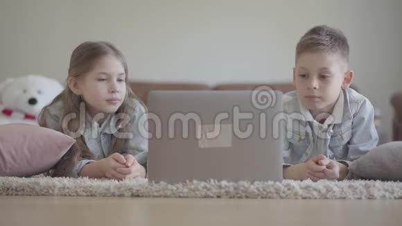 可爱的双胞胎男孩和女孩躺在地毯上看着电脑上有趣的东西笑着跳舞视频的预览图