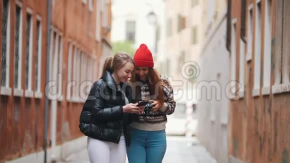 两位穿着温暖衣服的年轻女性旅行者拿着手机和相机在狭窄的街道上行走女人展示她的风趣视频的预览图