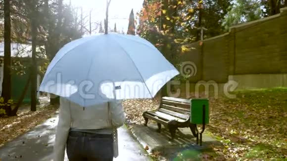 穿着白色皮夹克和牛仔裤的瘦女人在秋天的公园里带着蓝色的雨伞在雨中散步视频的预览图