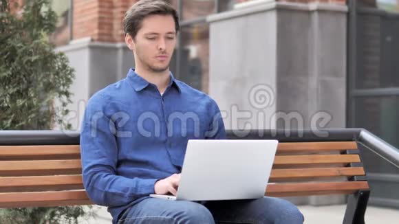 坐在户外脖子疼的年轻人在笔记本电脑上工作视频的预览图