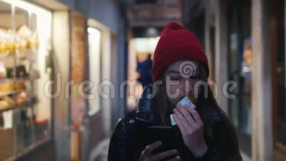 一位戴着红帽子的年轻女子在街上走着手里拿着电话一边吃着甜甜点视频的预览图