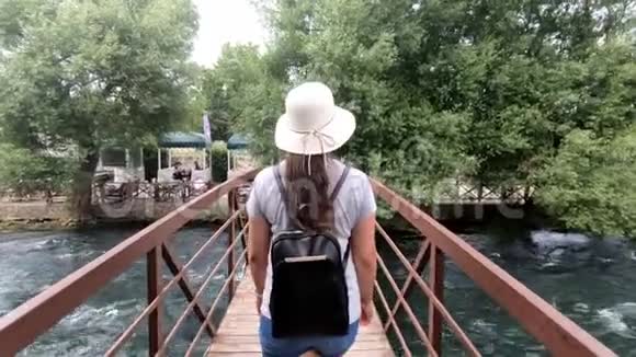 一位戴着帽子背着背包的旅游女孩正在河边的一座木桥上周围是许多绿色植物和树木视频的预览图