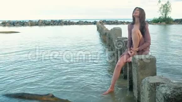 年轻的女人穿着衣服坐在石柱上站在海里做飞溅视频的预览图