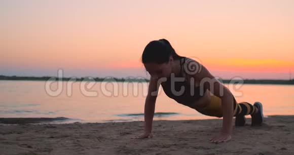 运动女孩在日出时在海滩上做俯卧撑穿着运动上衣和腿做俯卧撑的黝黑的年轻微笑女孩视频的预览图