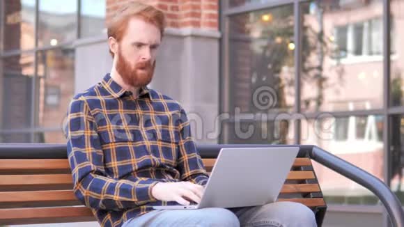 红发胡子青年男子在户外笔记本电脑工作时咳嗽视频的预览图