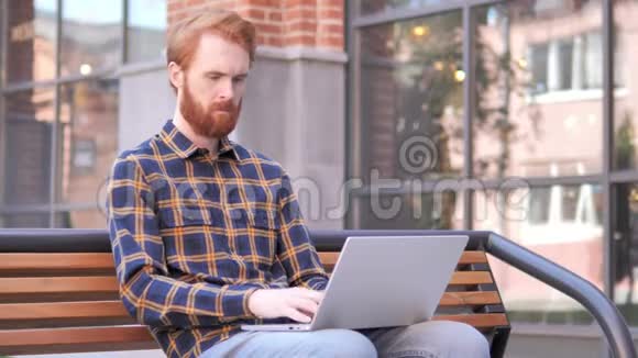 使用笔记本电脑坐在室外的红头发青年男子视频的预览图