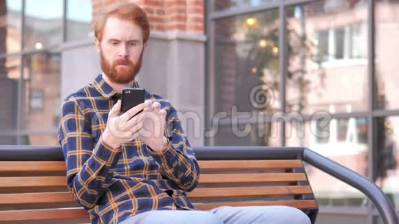 红胡子年轻人在智能手机上打字坐在户外长凳上视频的预览图