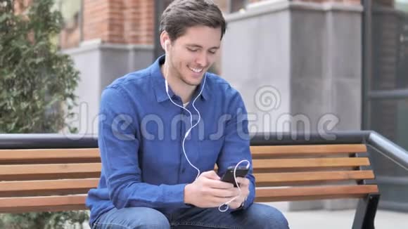 坐在长椅上听智能手机音乐的年轻人视频的预览图