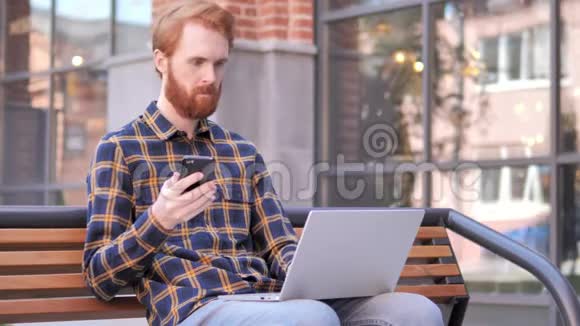 红胡子年轻人使用智能手机和笔记本电脑坐在长凳上视频的预览图