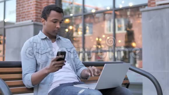 使用智能手机和笔记本电脑的非洲男人坐在长凳上视频的预览图