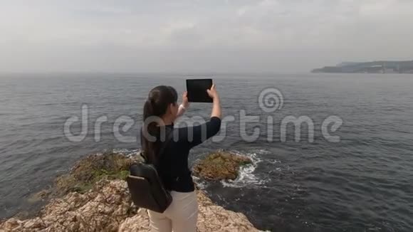 一个手里拿着平板电脑的女孩站在一片岩石海滩上拍摄着一幅美丽的照相机景色视频的预览图