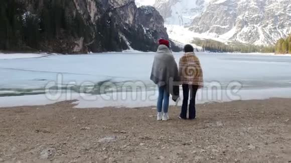 两个被毯子覆盖的年轻女人站在冰冻的海岸上看着群山视频的预览图