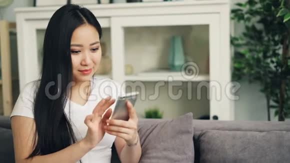 漂亮的亚洲女性穿着白色T恤正在使用智能手机触摸屏幕微笑和笑着查看社交媒体视频的预览图