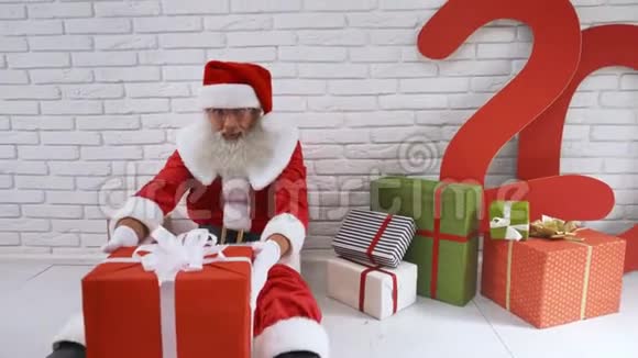 身穿圣诞老人服装的老人坐在墙边手里拿着礼品盒视频的预览图