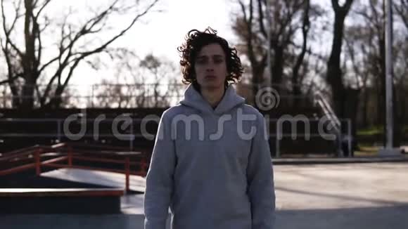 一个长着深色卷发的英俊年轻人的肖像穿着连帽衫的少年走过春天的溜冰公园视频的预览图