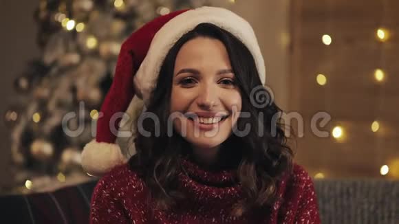 身着圣诞帽坐在圣诞树上闪闪发光的年轻美丽的布鲁内特女士的室内特写镜头视频的预览图