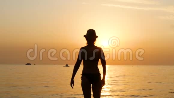 日落时分一位戴着帽子的年轻女子站在沙滩上穿着泳衣举起双手视频的预览图