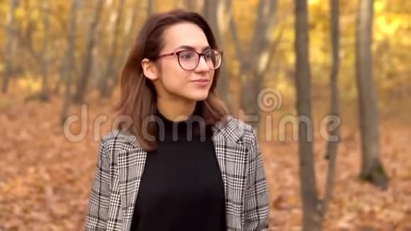 一个戴眼镜穿外套的年轻女子走在秋天的森林里黄色的叶子慢动作视频的预览图
