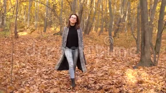 一个戴眼镜穿外套的年轻女子走在秋天的森林里黄色的叶子慢动作视频的预览图